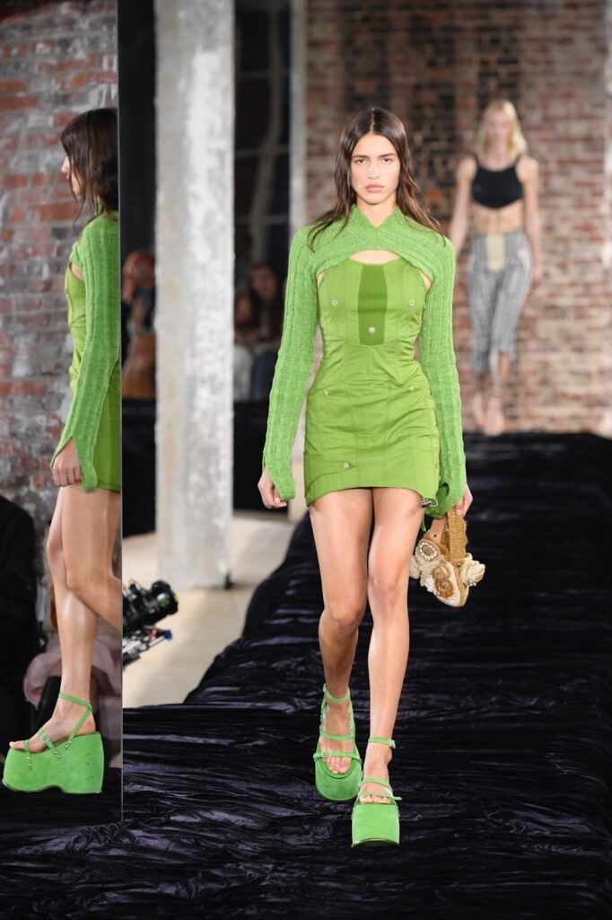 Moteris, dėvinti trumpą, prigludusią žalią suknelę ir avinti žalias basutes su platforma