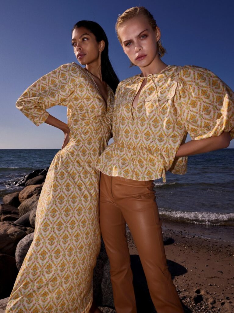 Dvi moterys: viena vilki ilgą, margą suknelę, kita - margą palaidinę pūstomis rankovėmis ir rudas odines kelnes.
