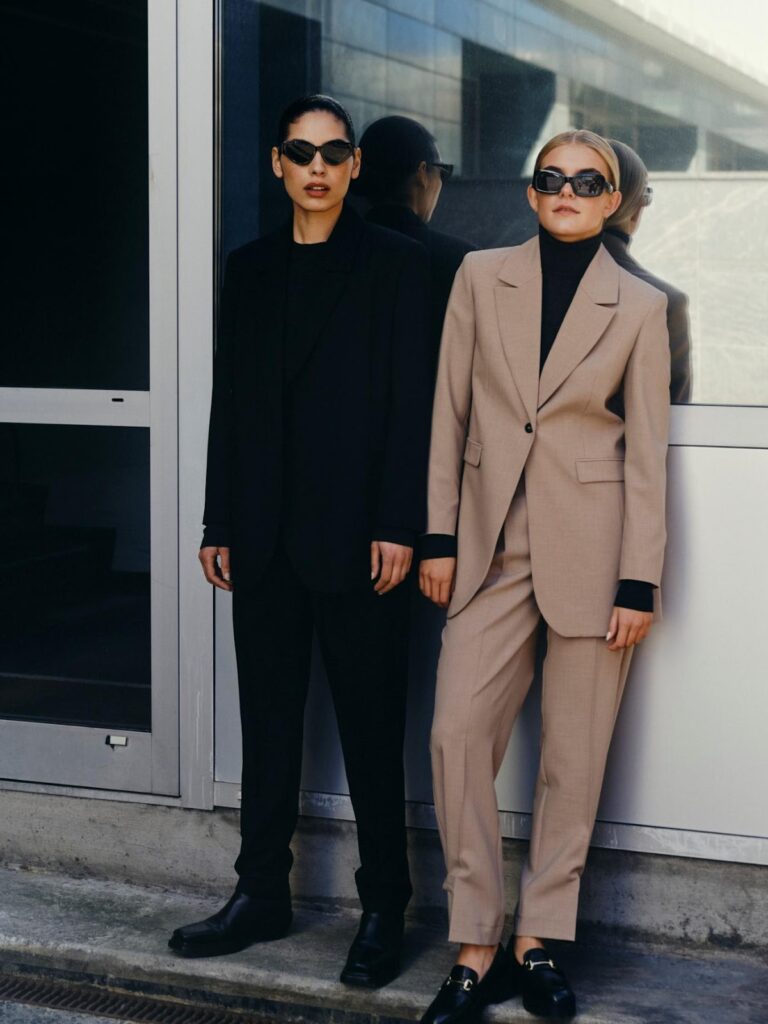 Dvi moterys - viena vilki juodą kostiumėlį, kita - smėlio spalvos kostiumėlį