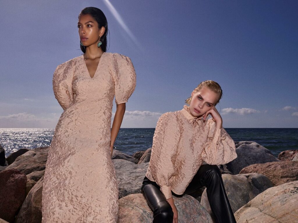 Dvi moterys: viena vilki smėlio spalvos suknelę pūstomis rankovėmis, kita - smėlio spalvos palaidinę pūstomis rankovėmis ir juodas odines kelnes.