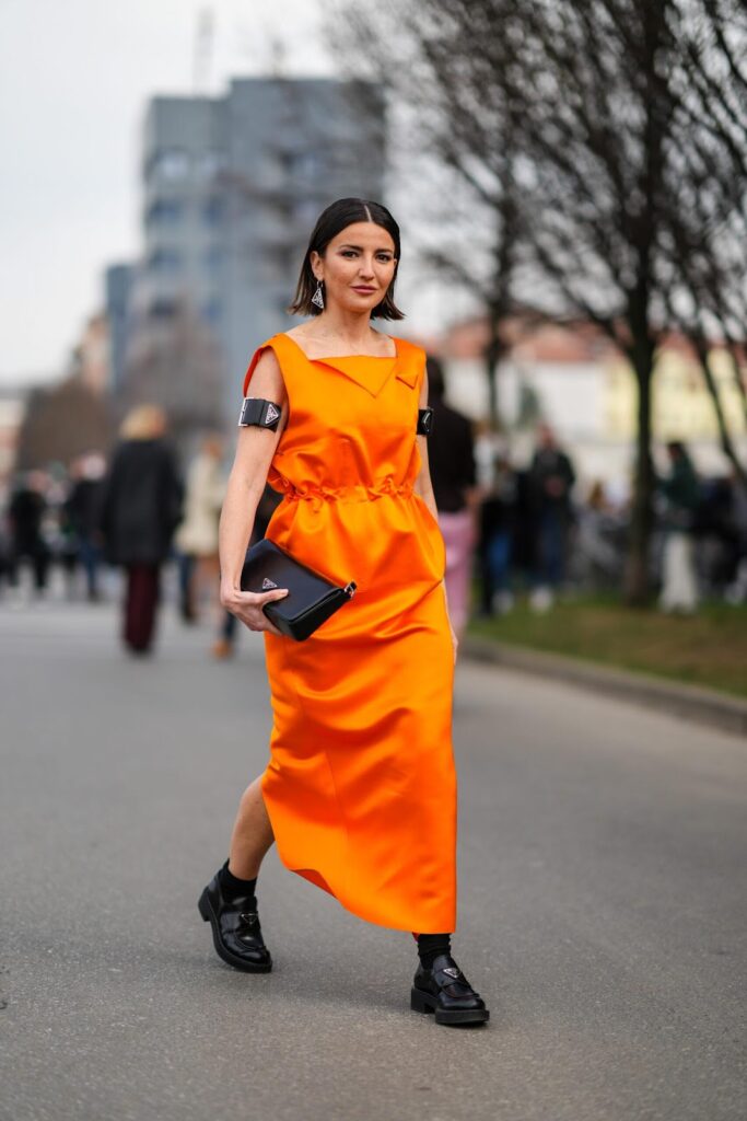 Moteris, dėvinti ryškiai oranžinę suknelę ir laikanti juodą delninukę