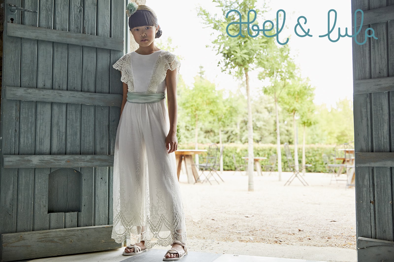 Maža mergaitė, dėvinti ilga balta nėriniuota suknele ir baltais sandalais