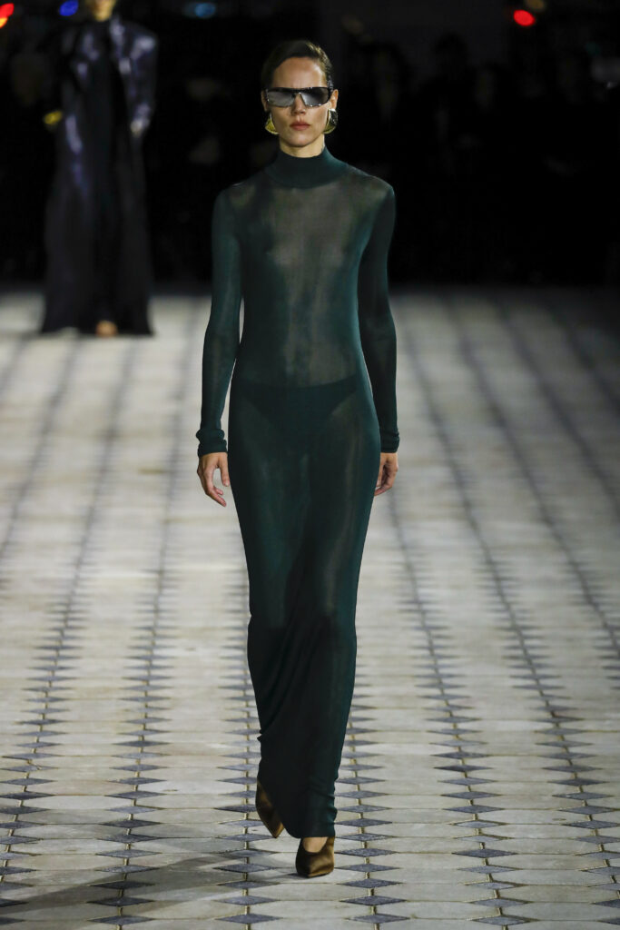 Podiumu žengiantis modelis, apsivilkęs ilgą, tamsiai žalią, permatomą suknelę