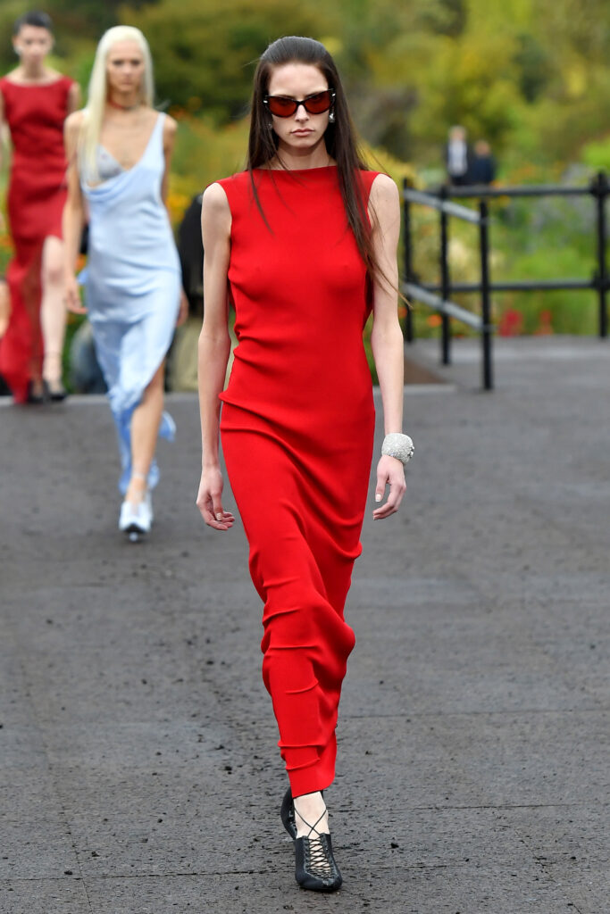 Podiumu žengiantis modelis, apsivilkęs ilgą raudoną suknelę